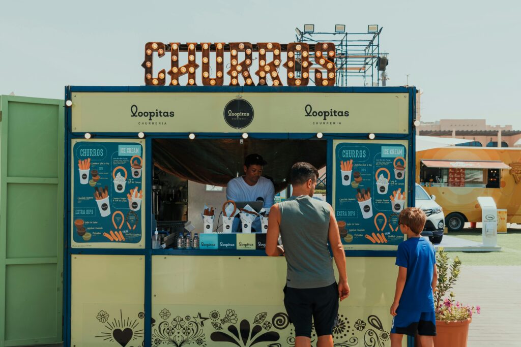 Father and son ordering churros at an outdoor market stand in sunny Dubai.